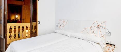 Habitación de hotel moderna con cama, ropa de cama blanca, una obra de arte geométrica en la pared y una puerta visible que conduce a otra habitación.