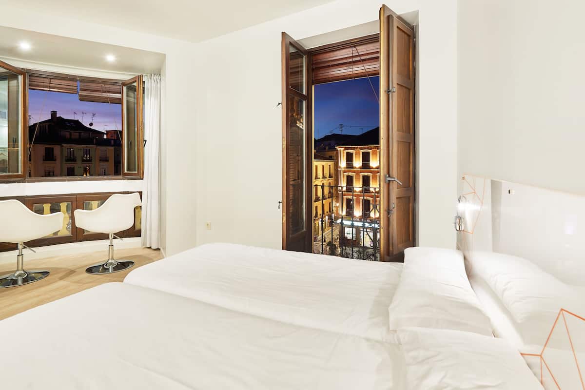 A hotel room with a large bed, two white chairs, a wooden door leading to a balcony, and a view of buildings at dusk.