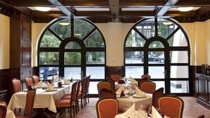Restaurante com várias mesas preparadas para refeições, grandes janelas em arco e vista para a rua.