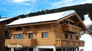 Una cabaña de madera con balcones, una pista de esquí al fondo y un letrero que dice "Le Grenier des Skis".