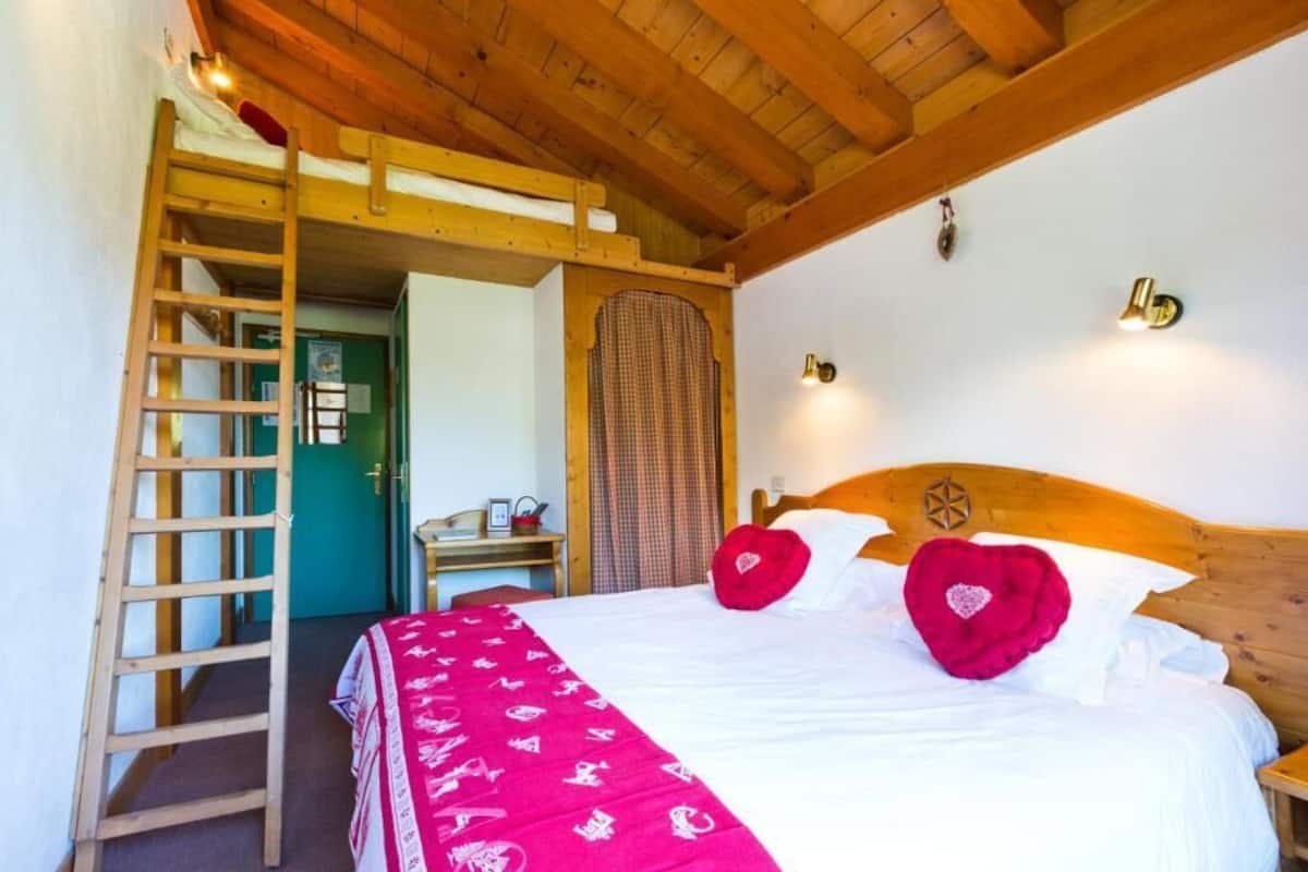 Une chambre avec un lit superposé, une échelle en bois, un lit avec du linge de lit blanc et des oreillers rouges, une tête de lit en bois et une petite table de chevet.
