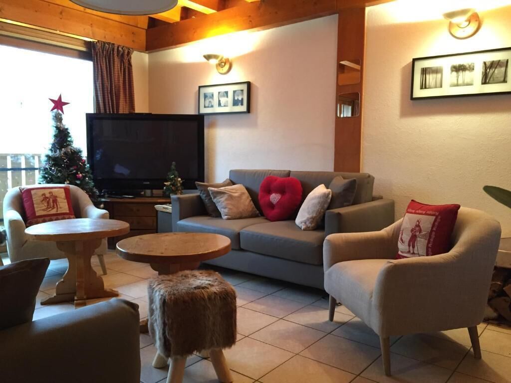 A cozy living room with a grey sofa, two armchairs, a round wooden coffee table, and a small ottoman. A television is mounted on the wall, and there are framed pictures above the sofa.