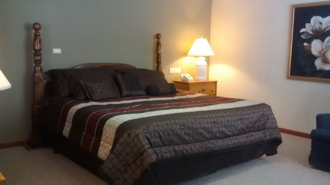 A bedroom with a bed, a nightstand with a lamp, a chair, and a painting on the wall.