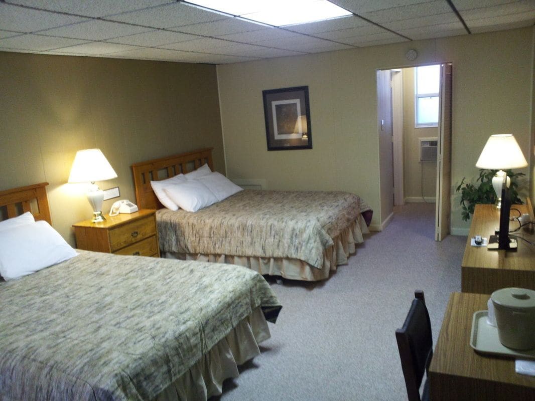A hotel room with two single beds, a desk with a lamp, a chair, and a small table with a cup.