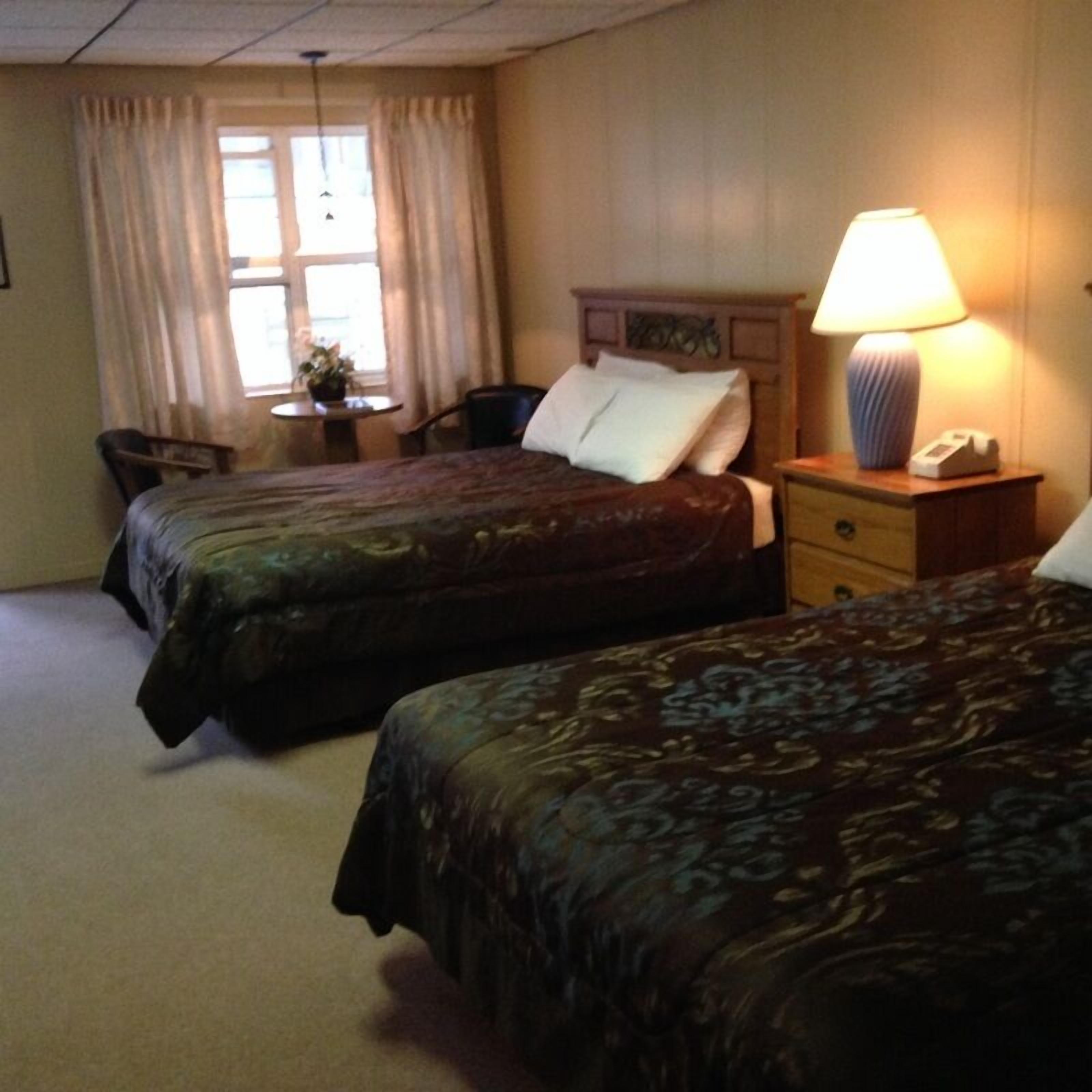 Quarto de hotel com duas camas, uma escrivaninha, uma cadeira e um abajur.