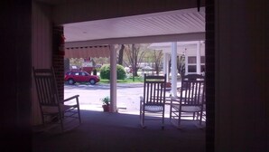 Une véranda avec deux fauteuils à bascule et un pot de fleurs, donnant sur une rue où une voiture rouge est garée devant un immeuble orné d’une enseigne.