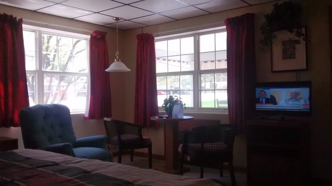 Ett hotellrum med en säng, två stolar, ett litet bord och en tv som visar en nyhetskanal.