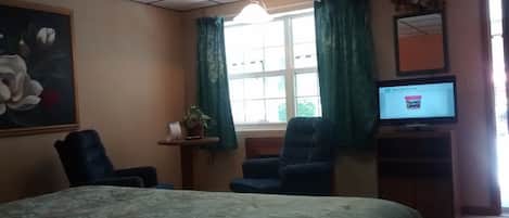 A hotel room with a bed, two armchairs, a small table with a plant, a television, and a window with curtains.