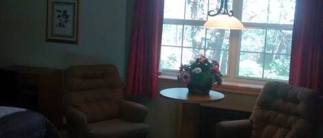 Una habitación con dos sillones, una mesita con un florero, una ventana con cortinas y un cuadro en la pared.