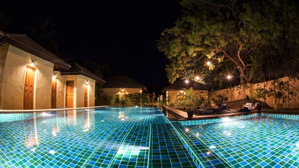 Una piscina iluminada en la noche, con piso de baldosas, rodeada de edificios con puertas y ventanas iluminadas, y árboles al fondo.