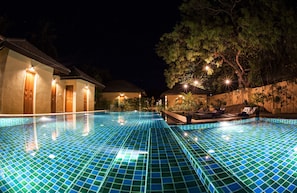 A nighttime swimming pool with tiled flooring, surrounded by buildings with illuminated doors and windows, and trees in the background. - The Spice Trail (Arugam Bay)