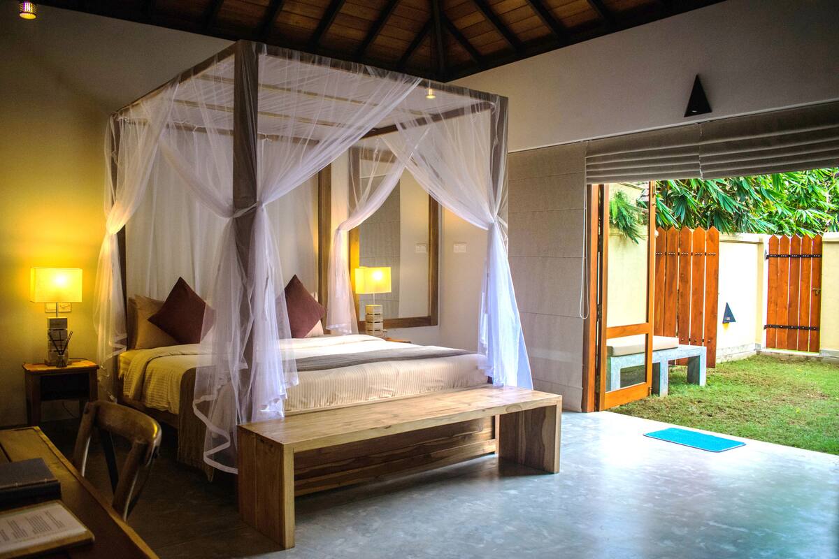 Una cama con dosel y cuatro columnas, lámparas de noche, un banco de madera y una mesita con un libro.