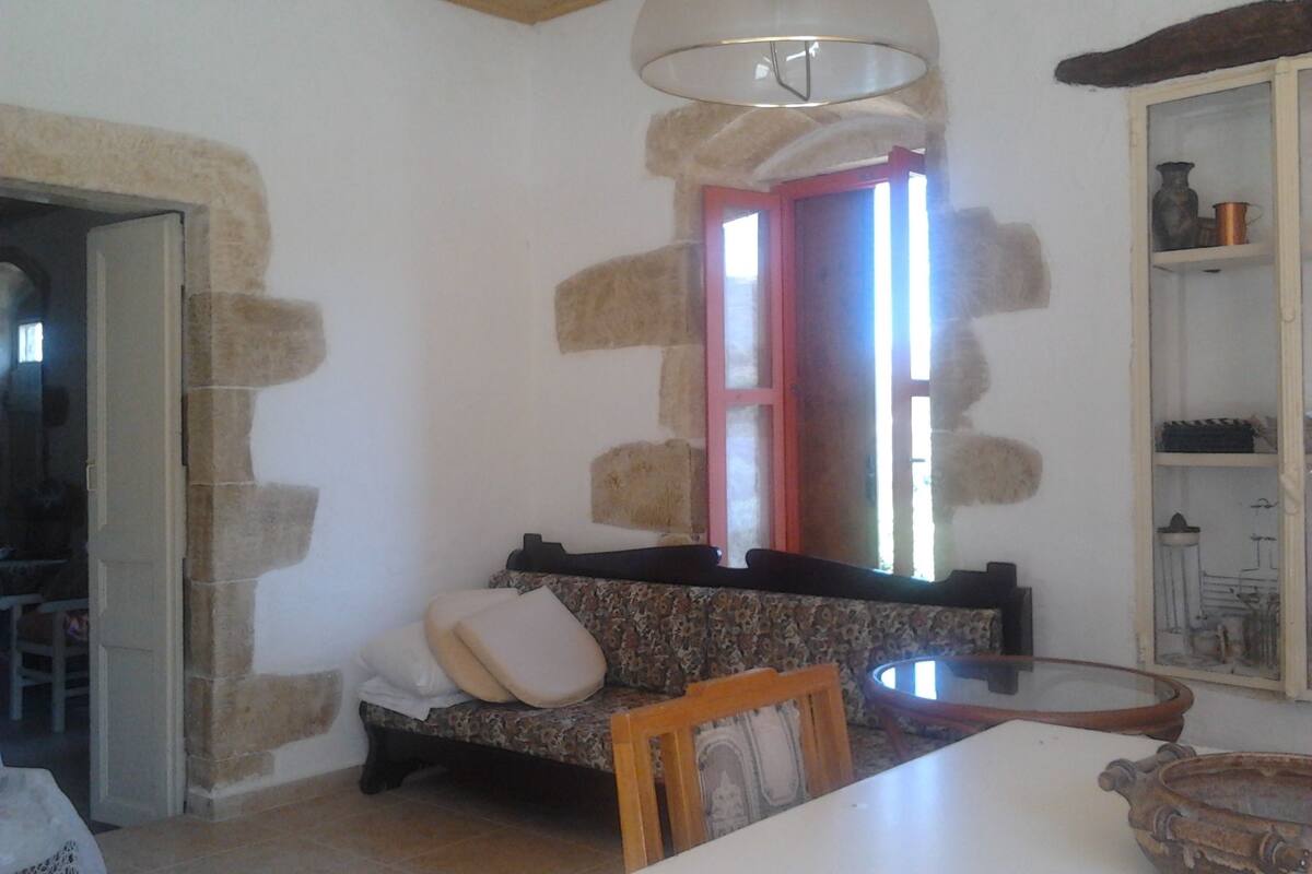 Una habitación con pared de piedra, un banco de madera, una mesa y una estantería con diversos objetos.