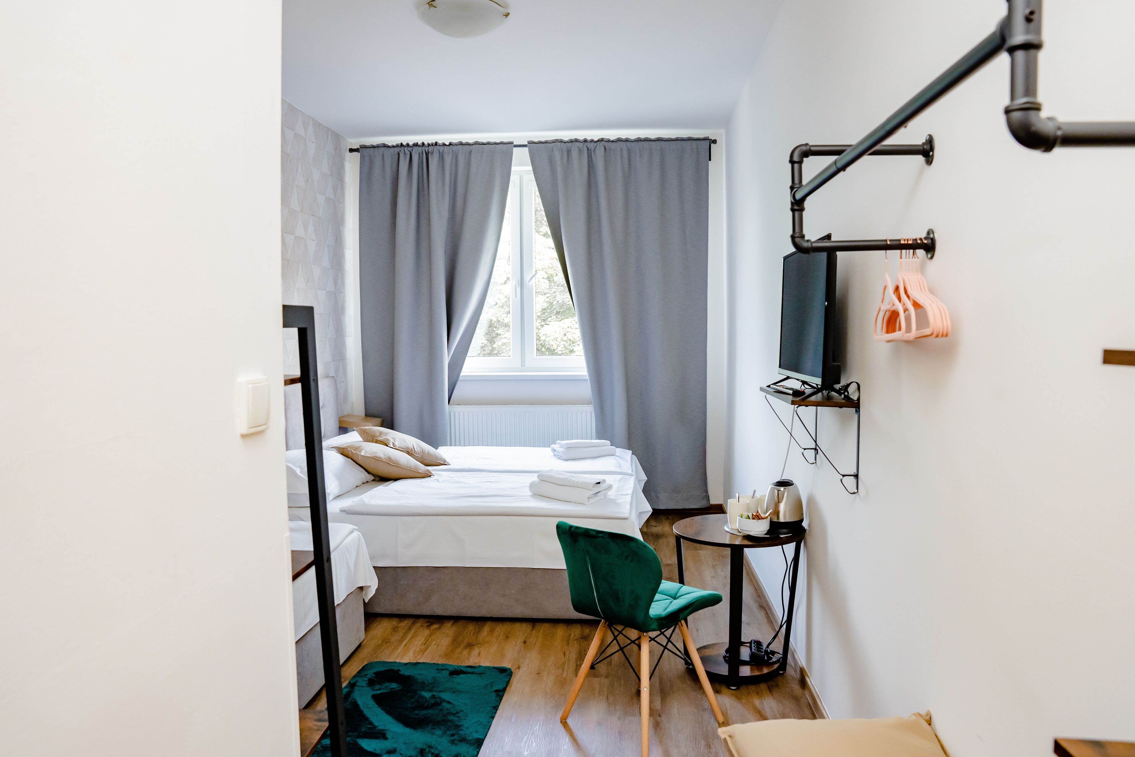 A modern hotel room with a bed, a green chair, a small table with a kettle, and a window with curtains.