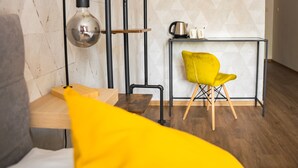 A modern room with a wooden table, a yellow chair, a metal shelf with a kettle and mugs, and a hanging light bulb. - Hotel Arko (Prague)