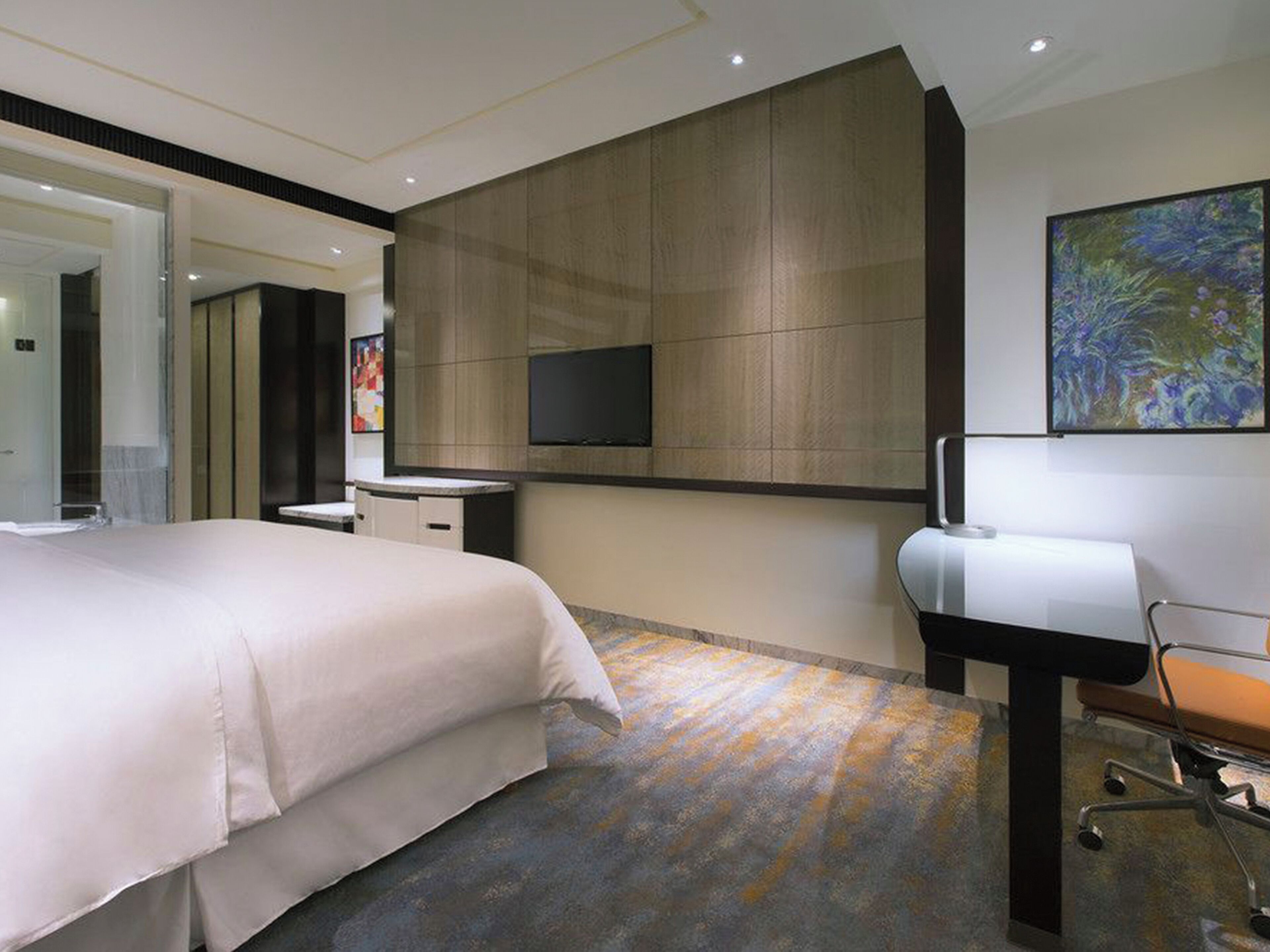 Habitació d'hotel moderna amb un llit gran, una taula de treball amb una cadira, un televisor de pantalla plana fixat a la paret i un quadre abstracte de grans dimensions sobre la taula.