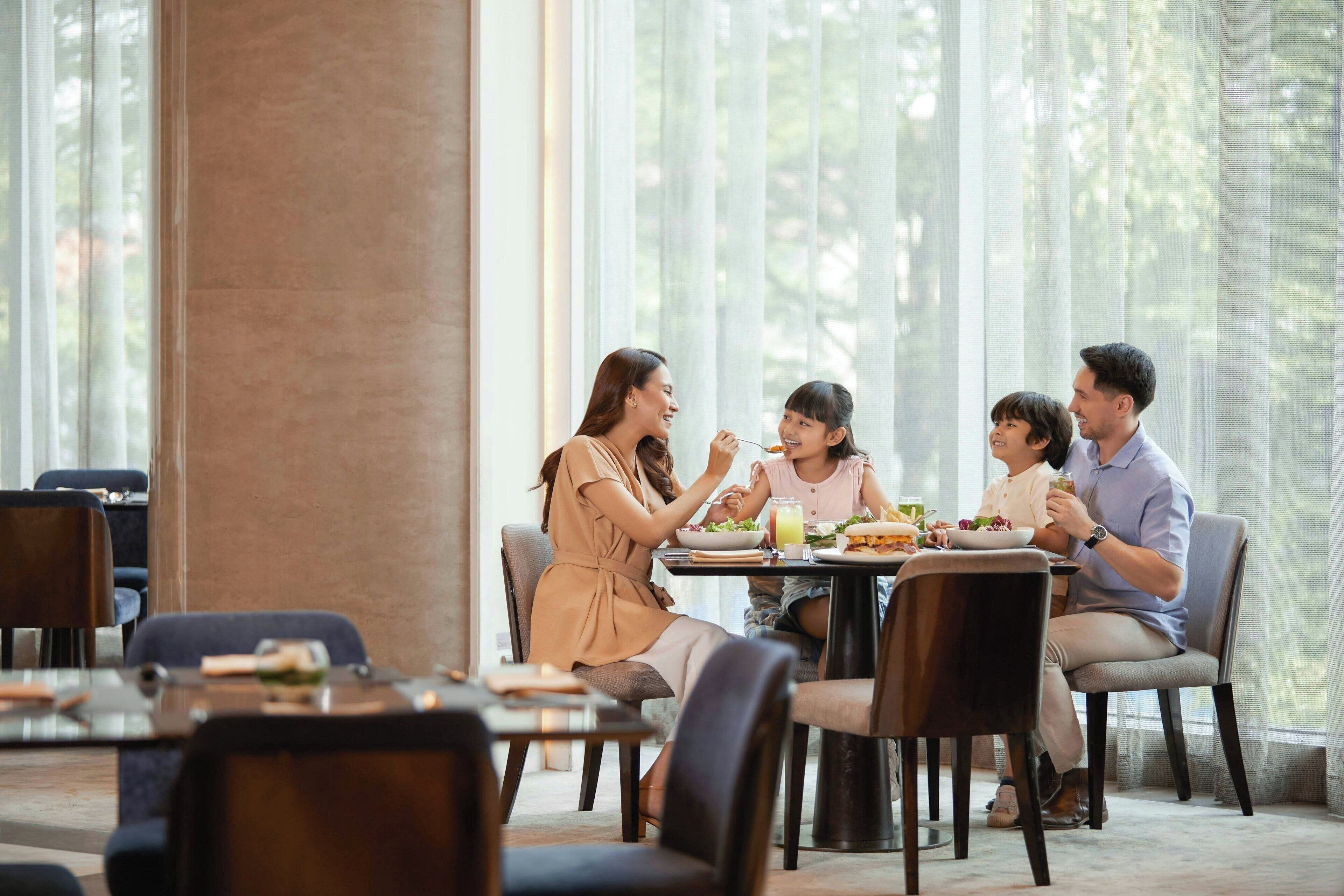 A family dining at a table in a well-lit room with large windows and a view of greenery outside.