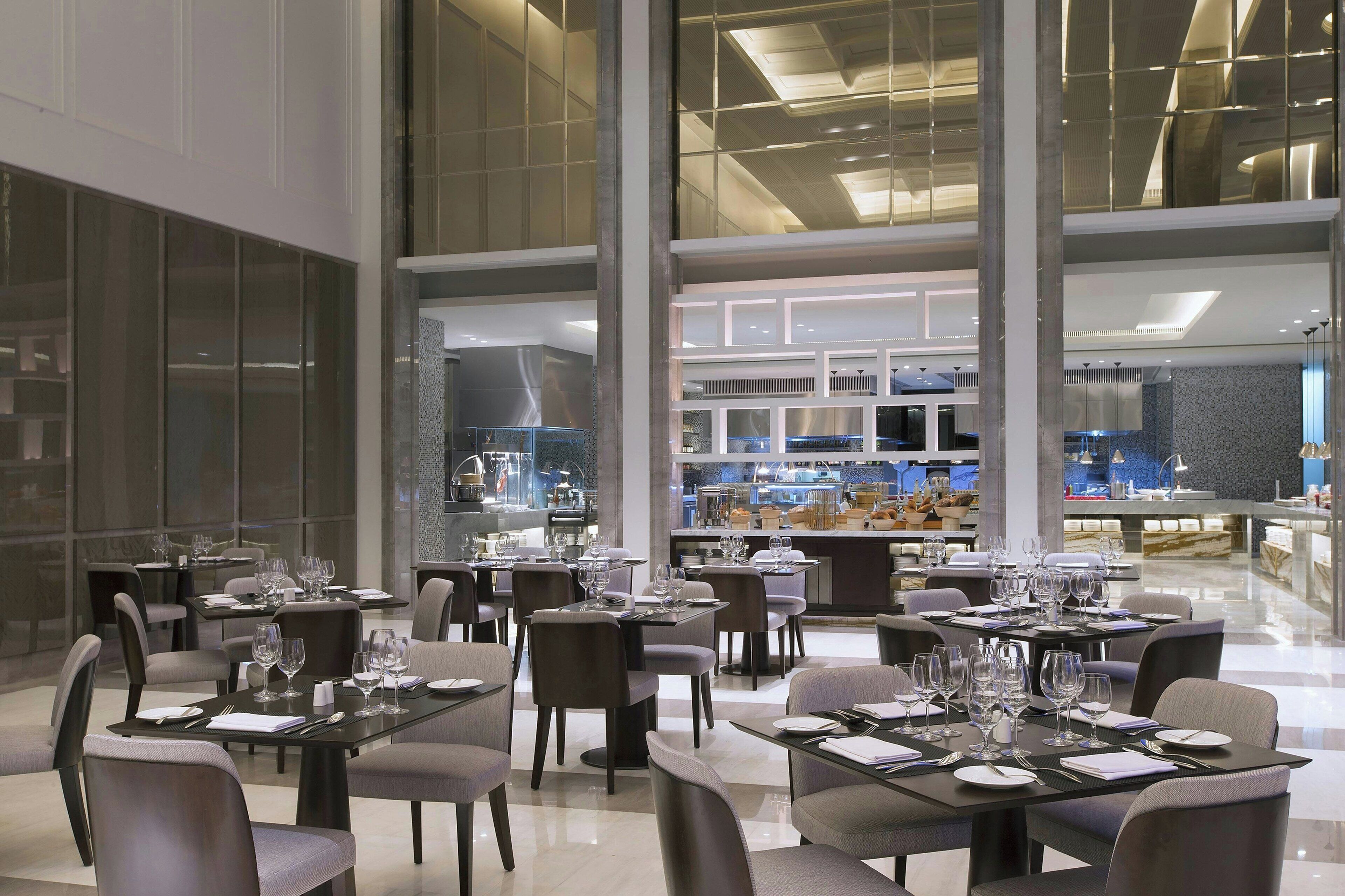 A modern dining area with multiple tables set for guests, large windows, and a view of the kitchen area.