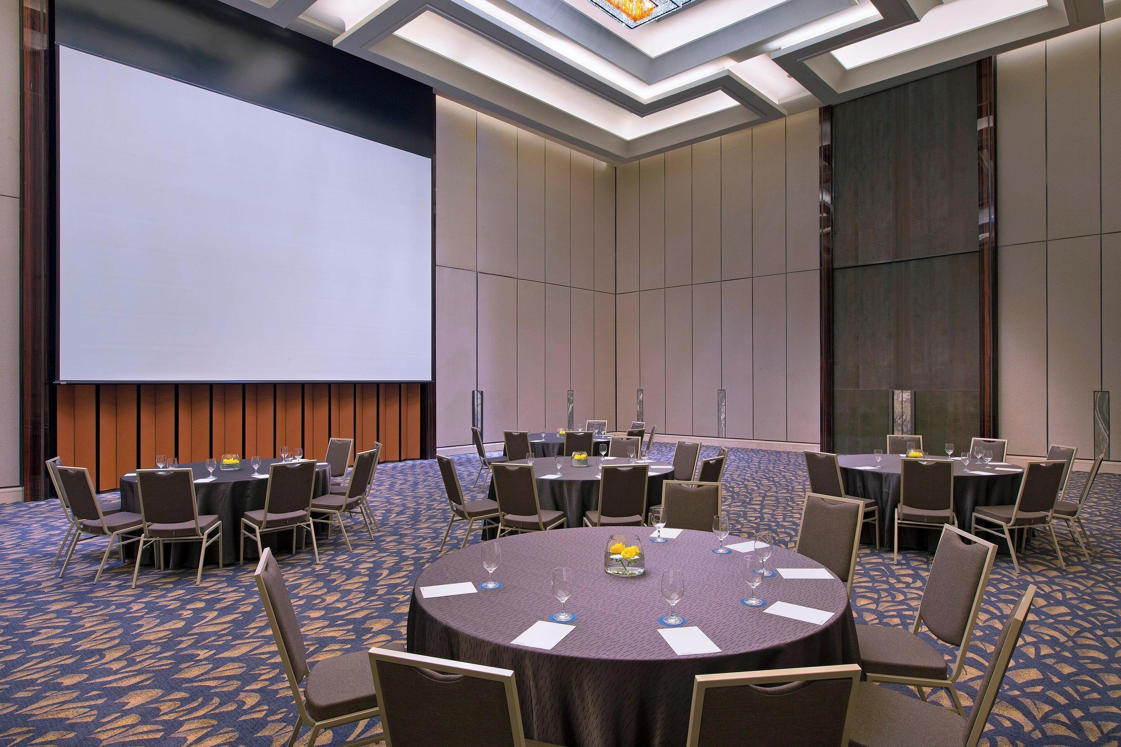 Un salón de conferencias con mesas redondas, sillas y una pantalla grande.