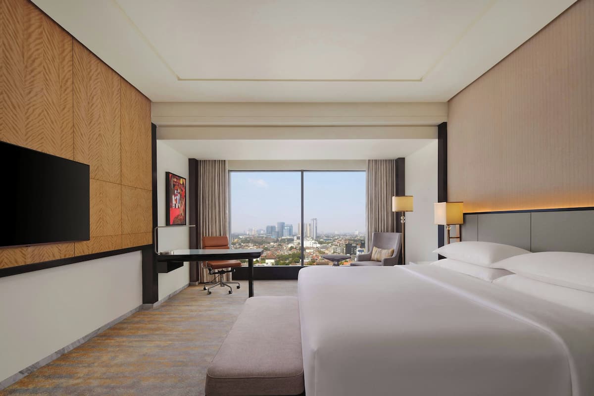 A hotel room with a large bed, a desk, a chair, a TV, and a view of the city.