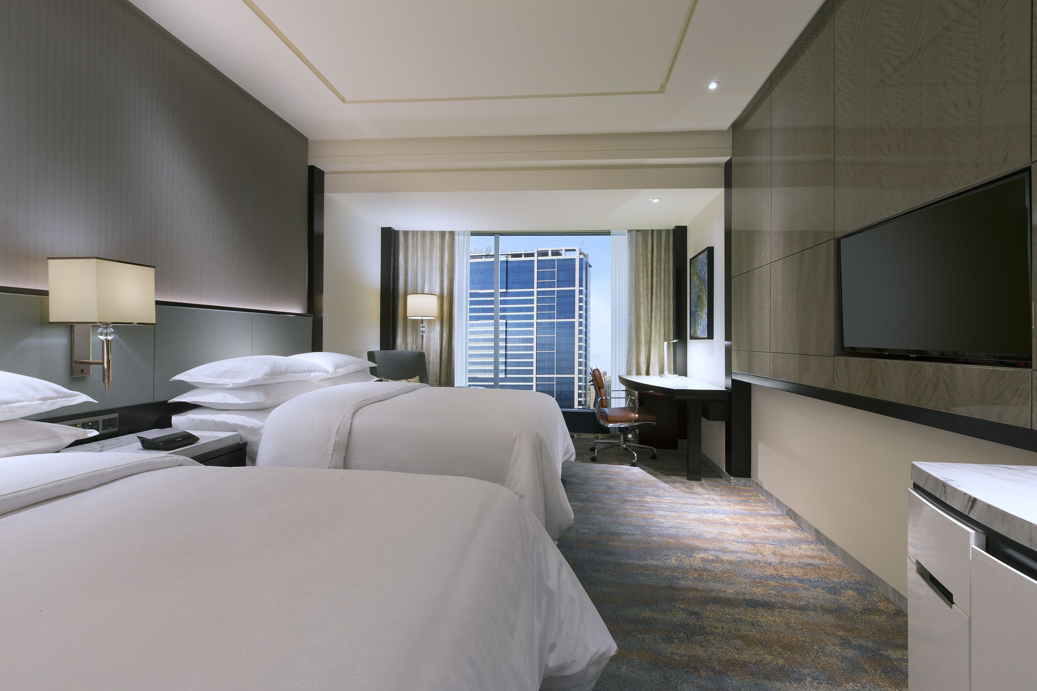 A hotel room with two beds, a desk, a chair, a TV, and a large window with a city view.