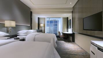 A hotel room with two beds, a desk, a chair, a TV, and a large window with a city view.
