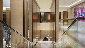 Eine moderne Hotelhalle mit Glastrennwand, einer zentralen Treppe und einem farbenfrohen, abstrakten Gemälde an der Wand.