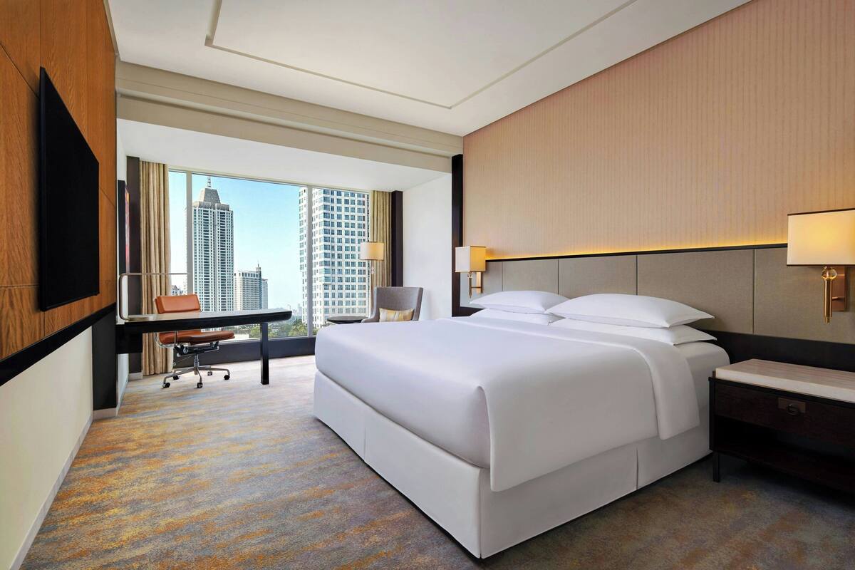 Une chambre d’hôtel avec un grand lit, un bureau avec une chaise, une télévision et une vue sur de hauts immeubles.