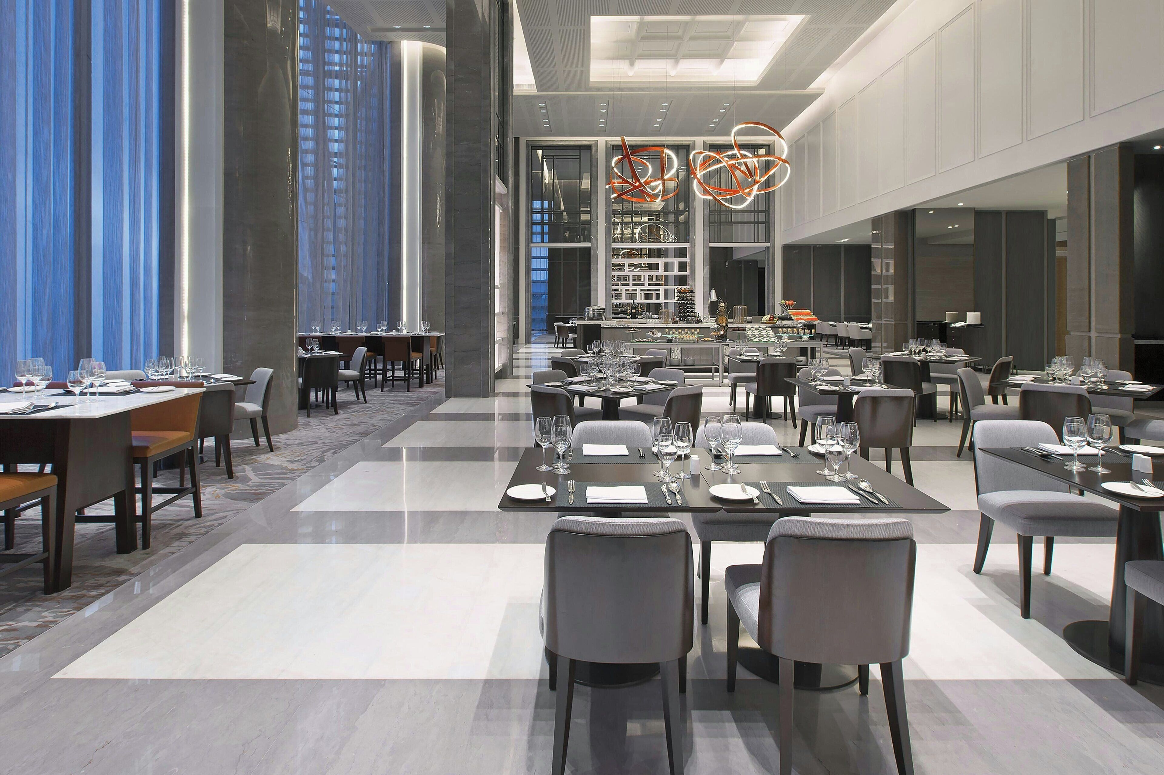 A spacious, modern dining area with multiple tables set for guests, featuring a high ceiling and large windows.