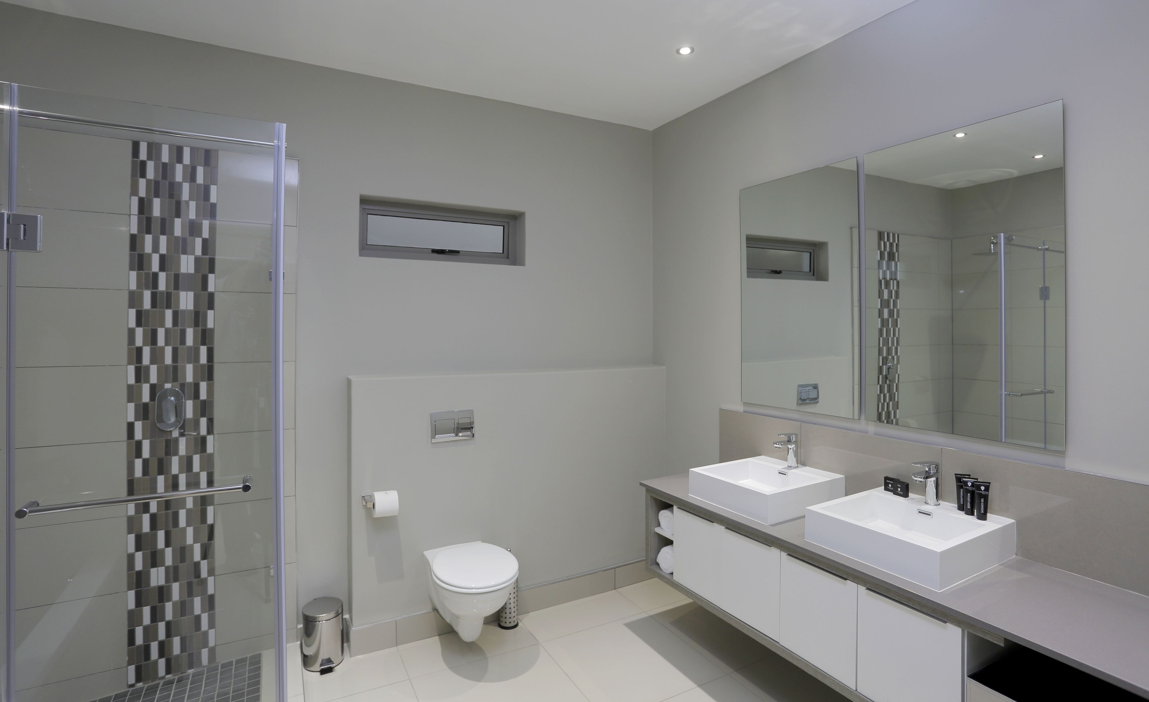 Un baño moderno con cabina de ducha de vidrio, inodoro y dos lavamanos con albornoces.
