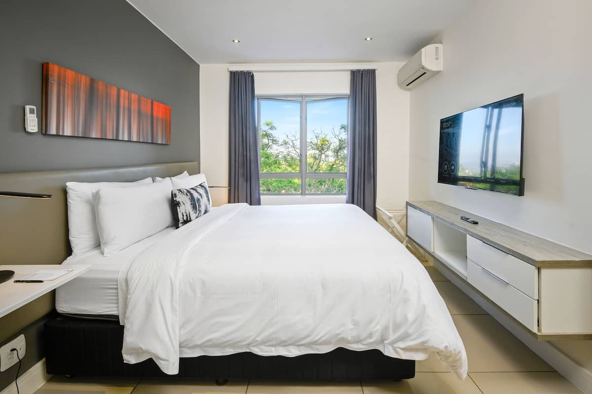 A modern hotel room with a large bed, a flat-screen TV, a desk with a chair, and a balcony with a view of greenery.