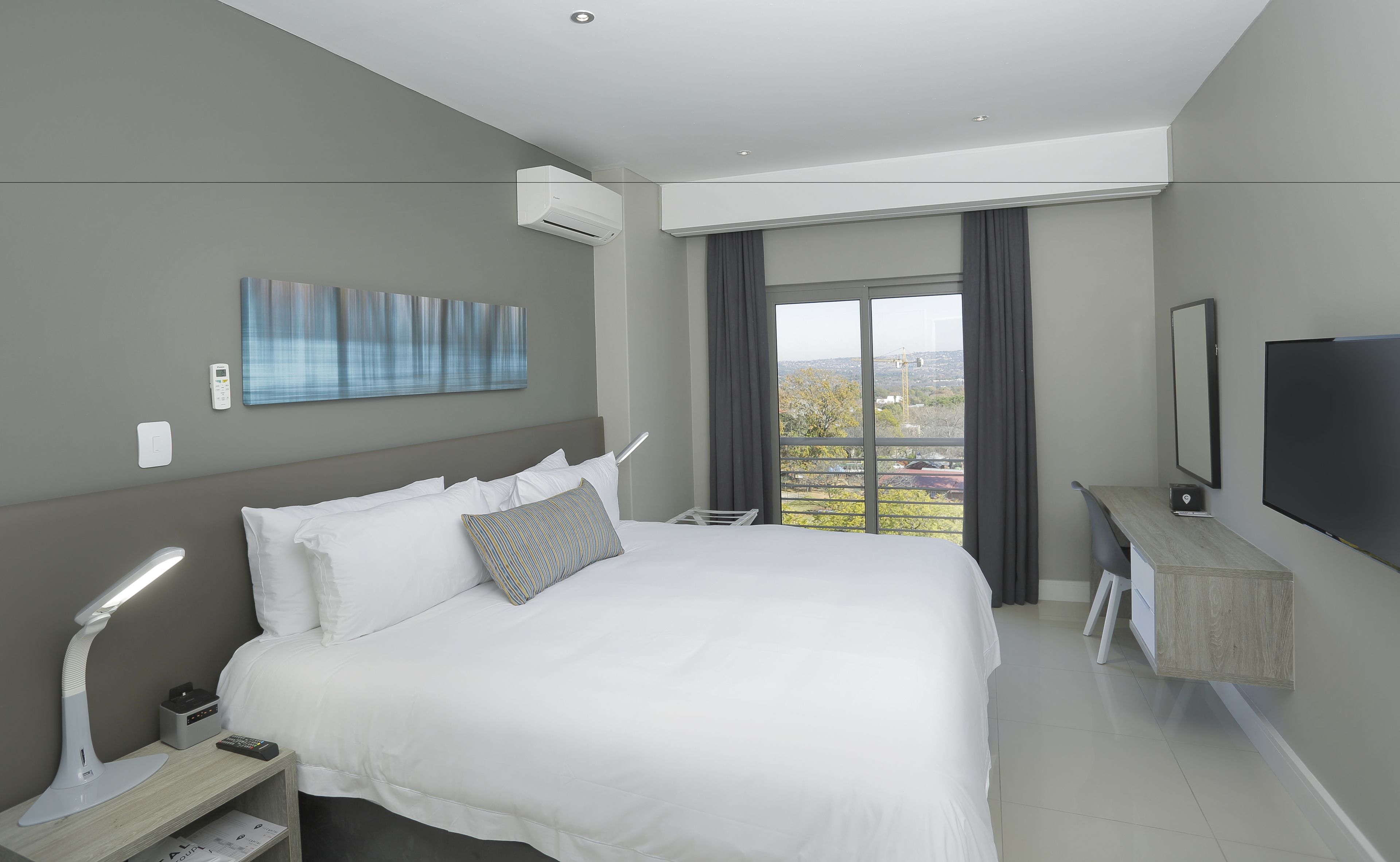 Habitación de hotel moderna con una cama grande, mesita de noche, lámpara y un televisor sobre un soporte de madera.