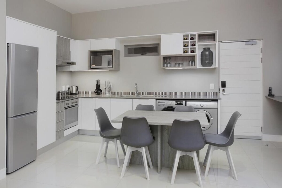 A modern kitchen with a built-in oven, microwave, and dishwasher, a dining area with a central island and four chairs, and white cabinetry.