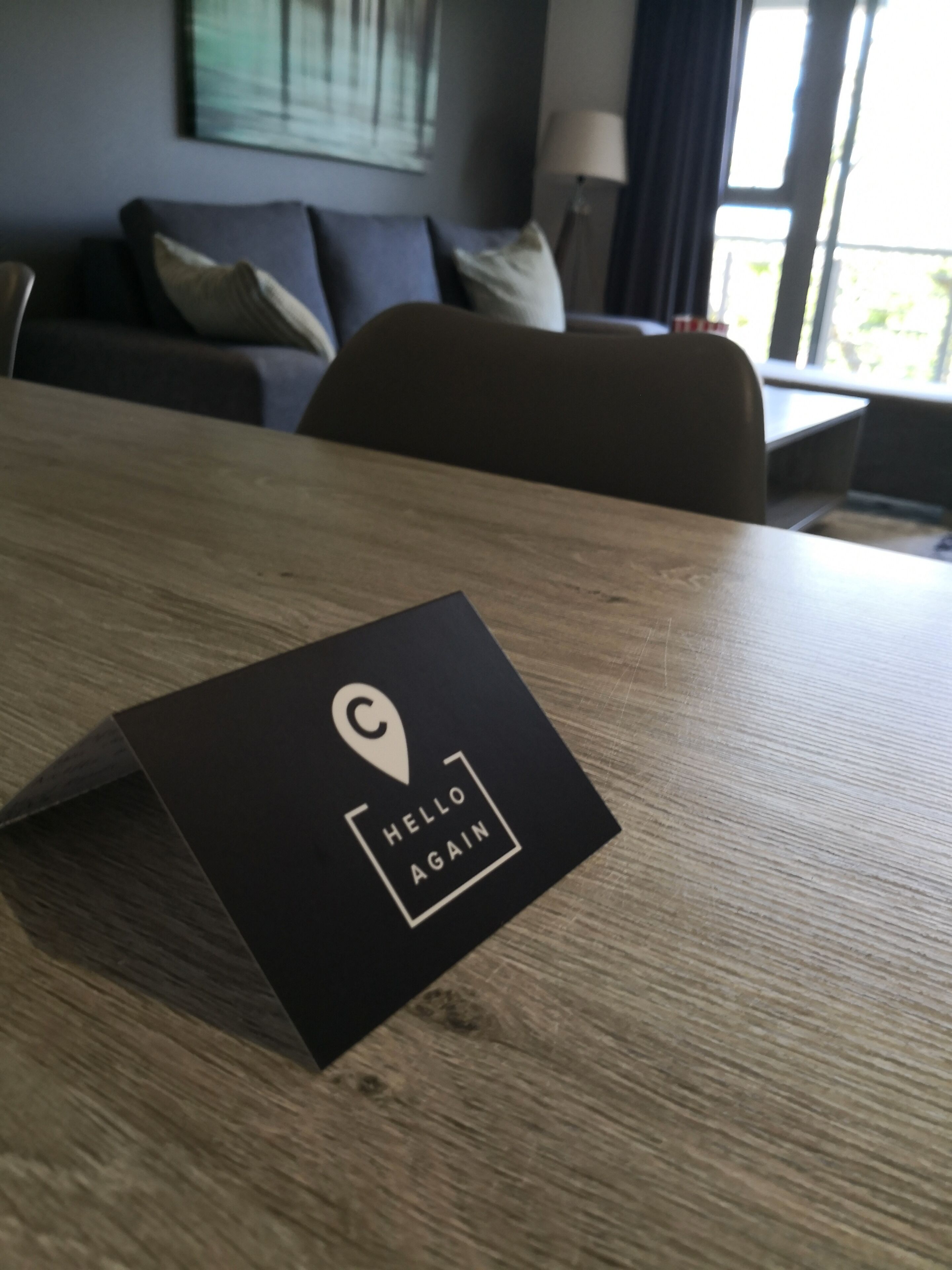A black card with a location pin icon and the text "HELLO AGAIN" on a wooden table in a room with a couch and a lamp in the background.