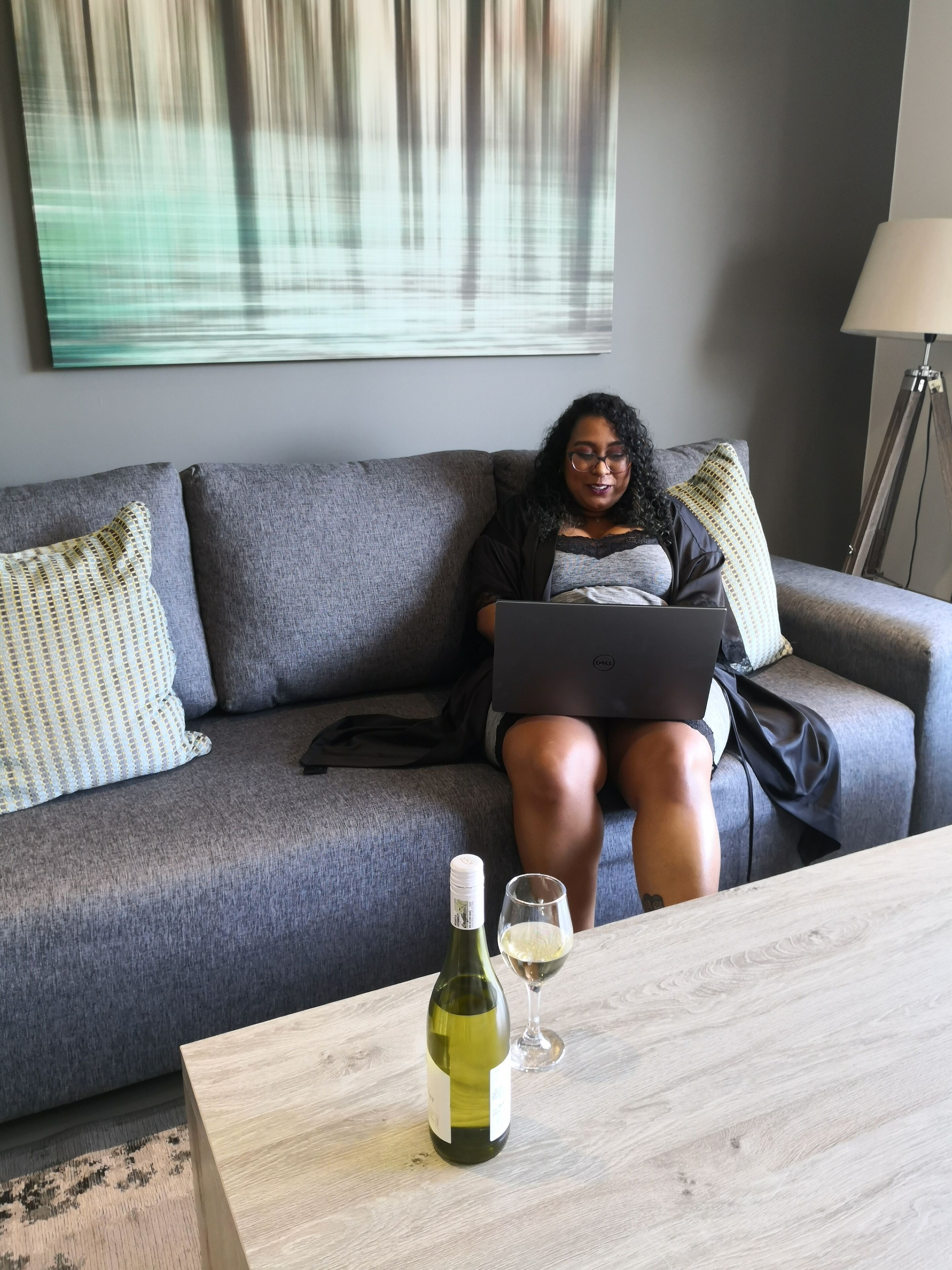 Eine Person sitzt auf einem grauen Sofa mit einem Laptop, einer Weinflasche und einem Glas auf einem Holztisch.