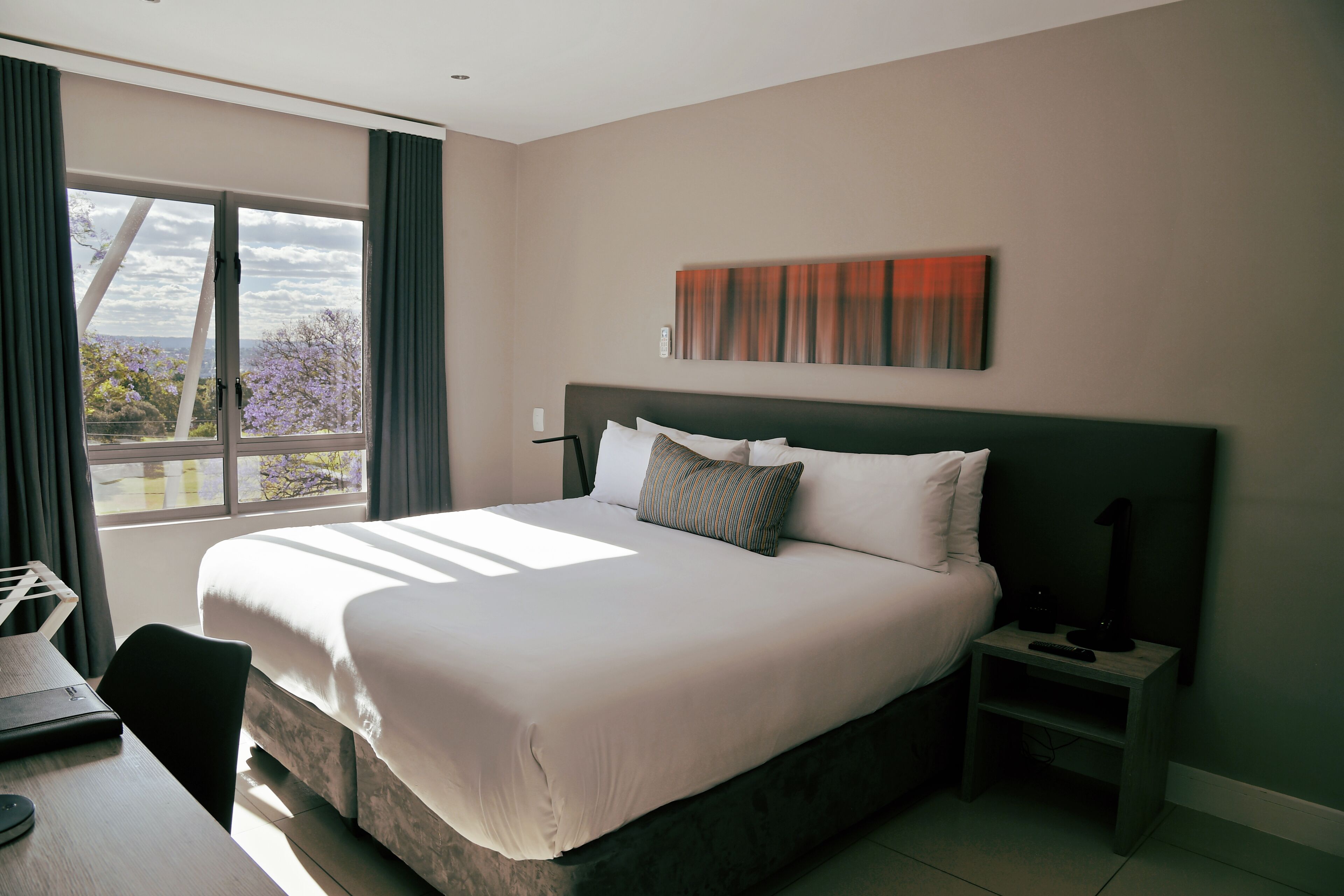 Una habitación de hotel moderna con una cama grande, una mesita de noche, un escritorio y un ventanal con cortinas.