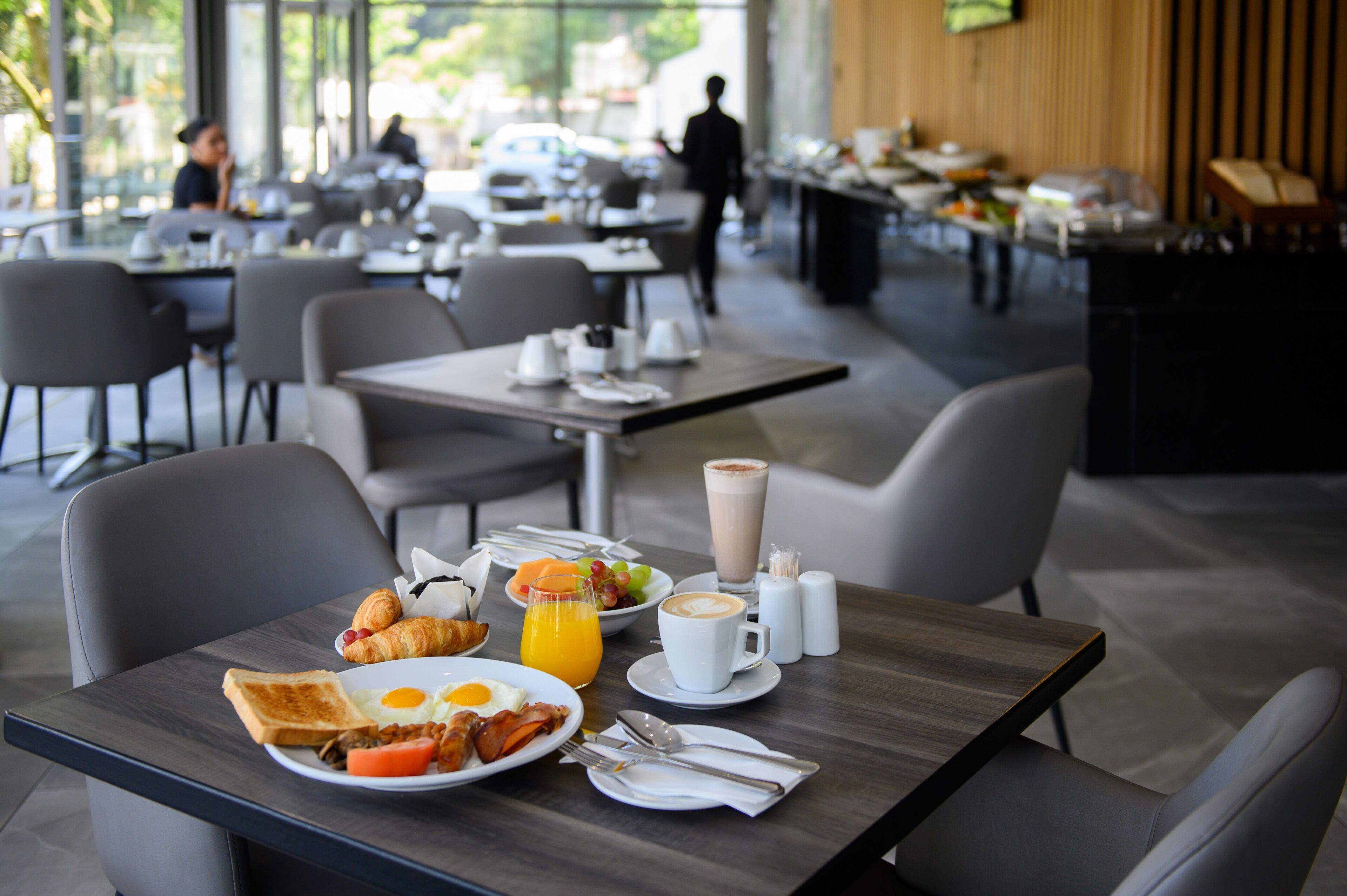 Desayuno con un plato de huevos, tostadas y croissants, un vaso de zumo de naranja y una taza de café sobre una mesa.
