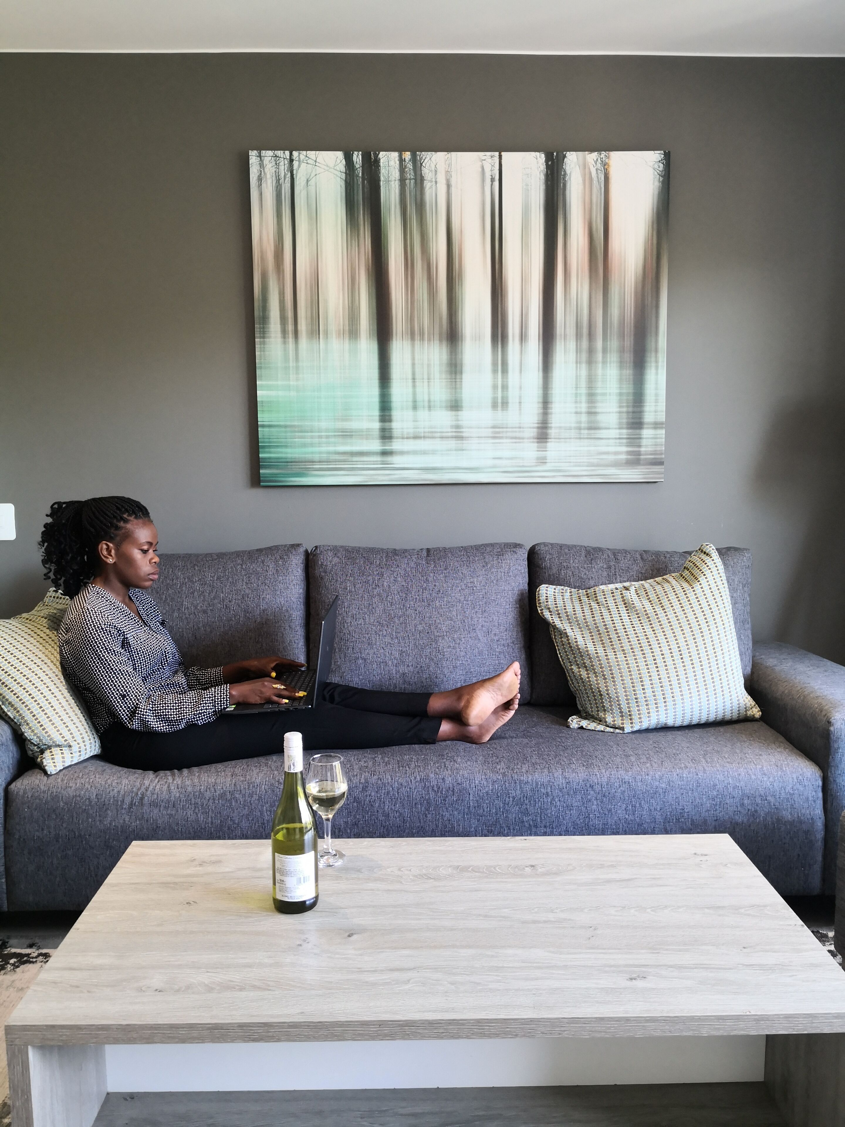 A person sitting on a grey sofa with a laptop, a bottle of wine, and a glass on a wooden coffee table.