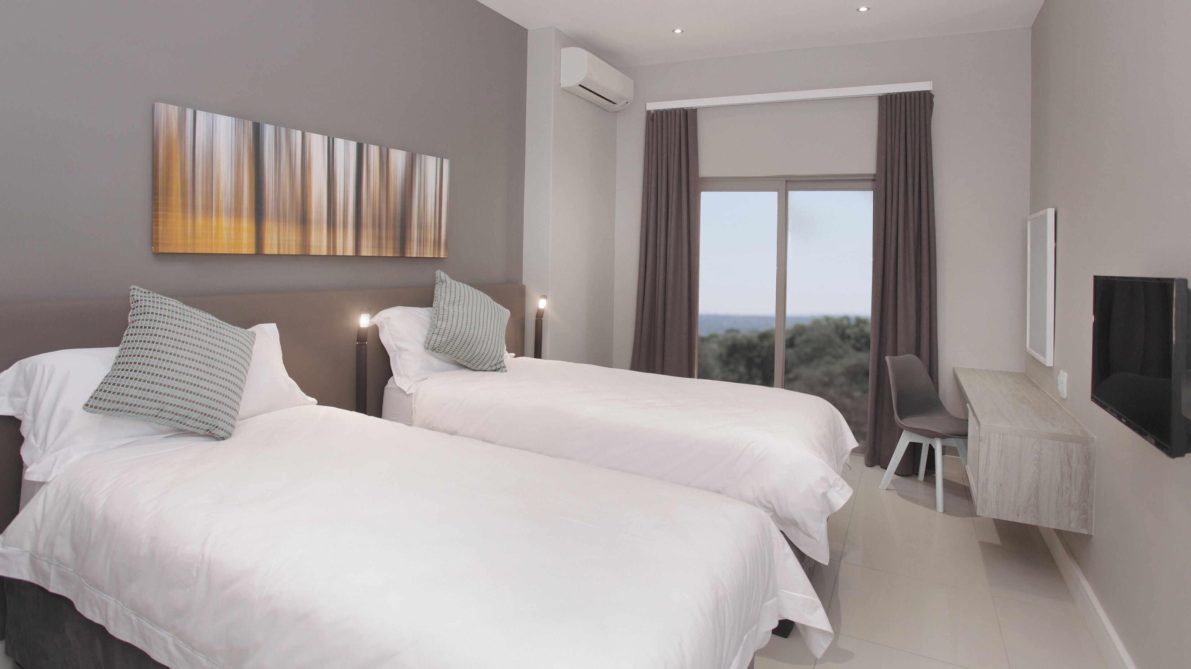 A hotel room with two beds, a desk, a TV, and a large window with curtains.