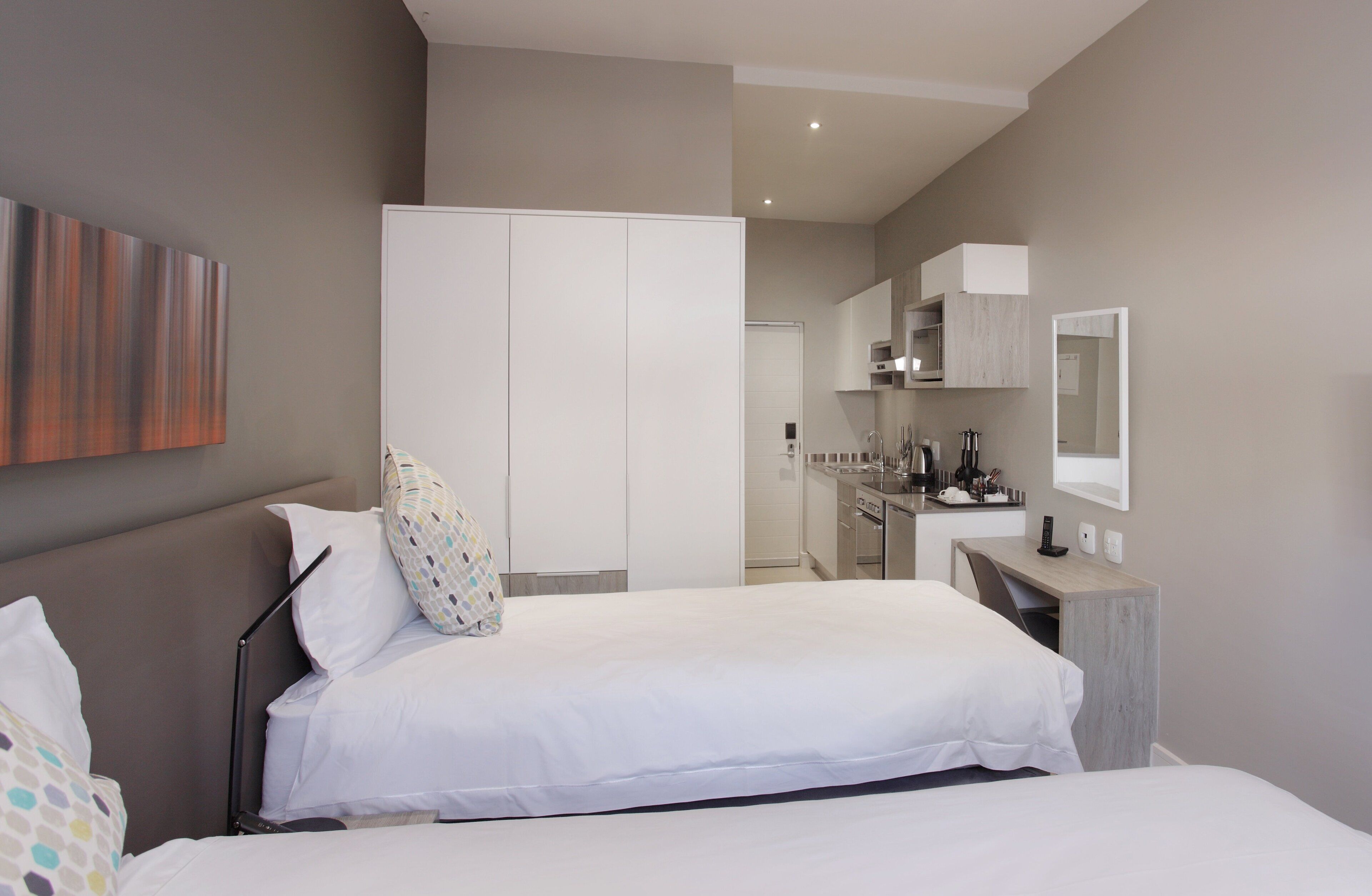 A modern hotel room with a bed, bedside table, and a small desk with a chair. There is a kitchenette in the background.
