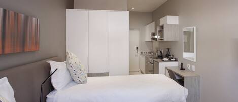 A modern hotel room with a bed, bedside table, and a small desk with a chair. There is a kitchenette in the background.