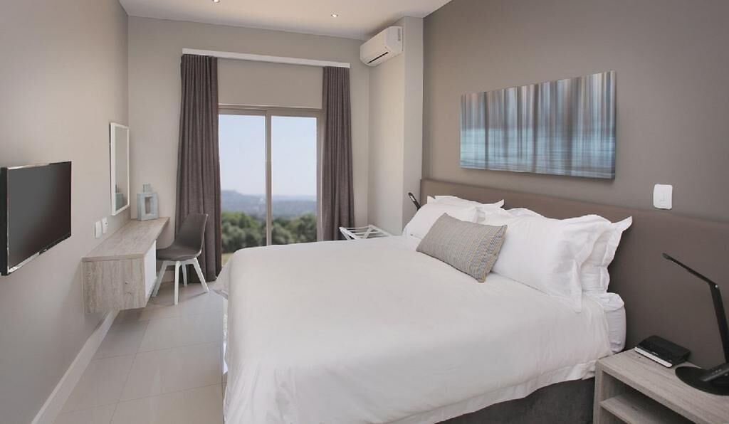 A modern hotel room with a large bed, a desk with a chair, a flat-screen TV, and a large window offering a view of the outdoors.