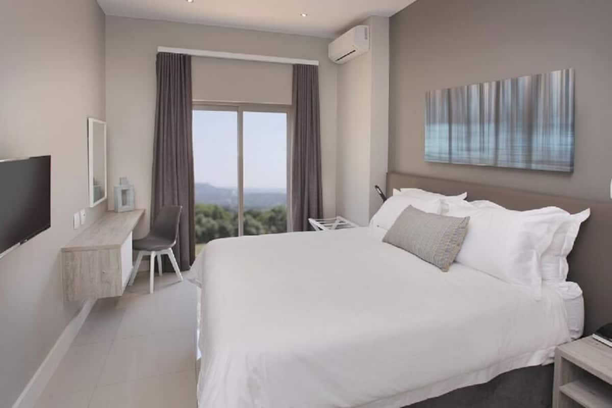 A modern hotel room with a large bed, a desk with a chair, a flat-screen TV, and a large window offering a view of the outdoors.