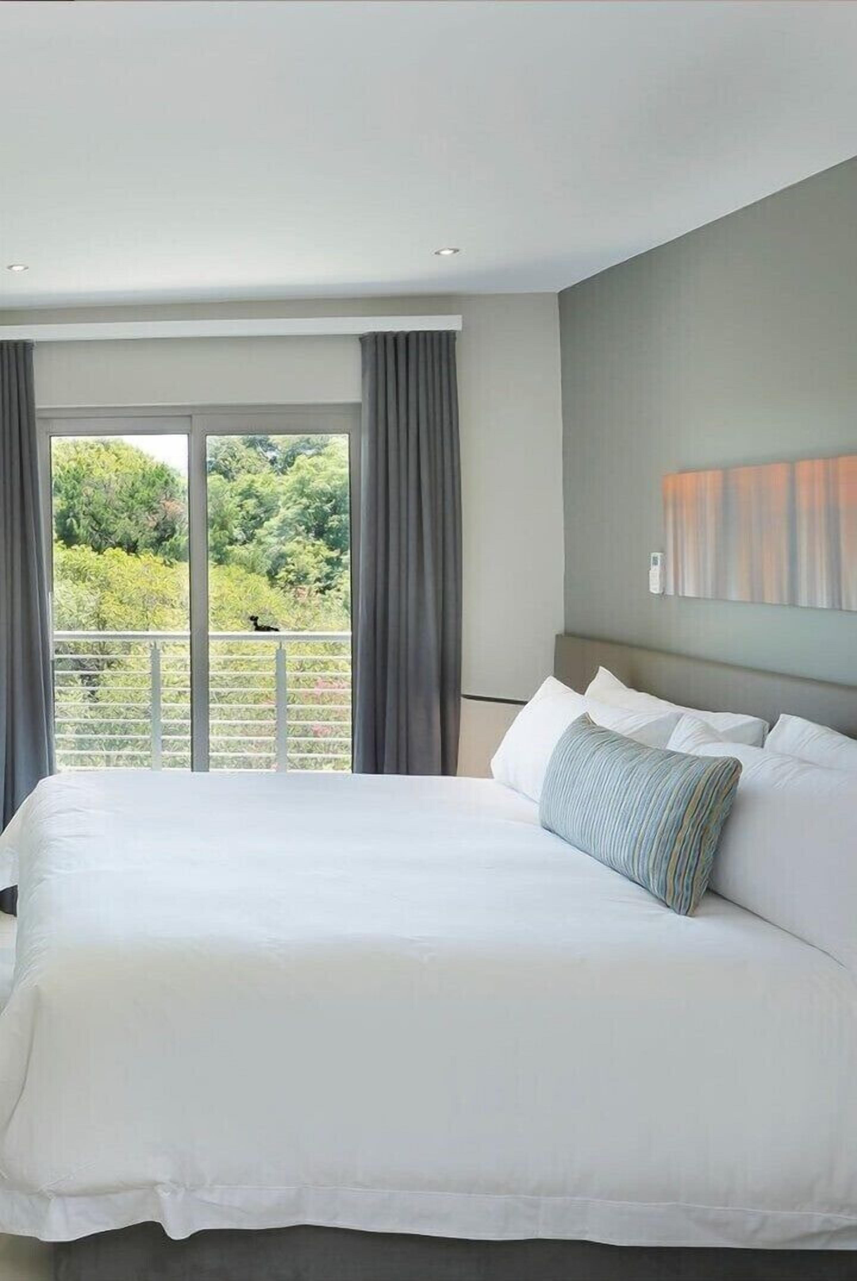 A hotel room with a large bed, a balcony with curtains, and a view of greenery.