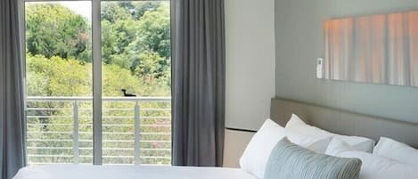 A hotel room with a large bed, a balcony with curtains, and a view of greenery.