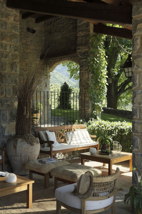 A rustic outdoor seating area with stone walls, a wooden bench, and a table with a vase and a pitcher.