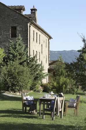 Un área de comedor al aire libre con un edificio de piedra al fondo, rodeada de vegetación.
