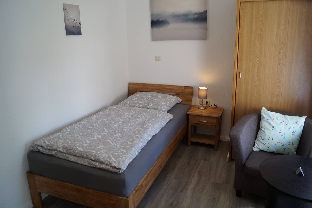 Un lit simple avec une tête de lit en bois, une table de chevet avec une lampe, un canapé gris avec un coussin à motifs floraux et une petite table ronde.