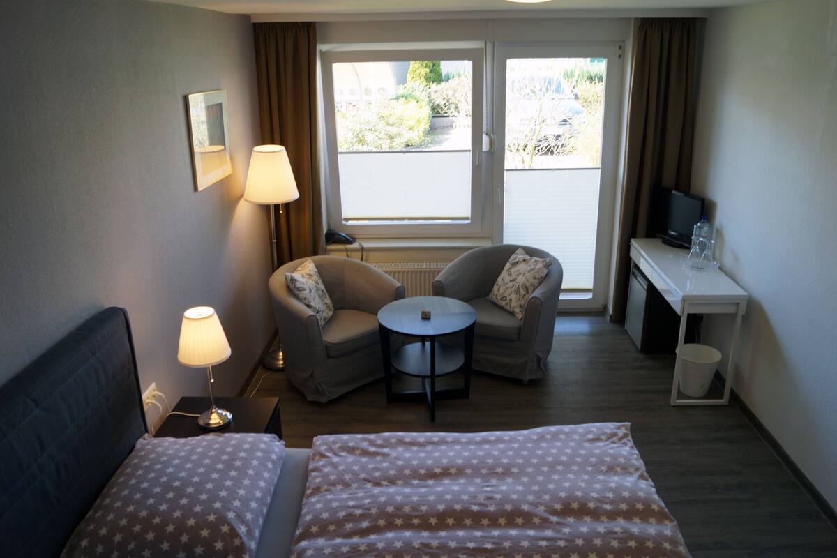 Une chambre d’hôtel comprenant un lit, deux fauteuils, une petite table ronde, un bureau avec une télévision et une fenêtre avec des rideaux.