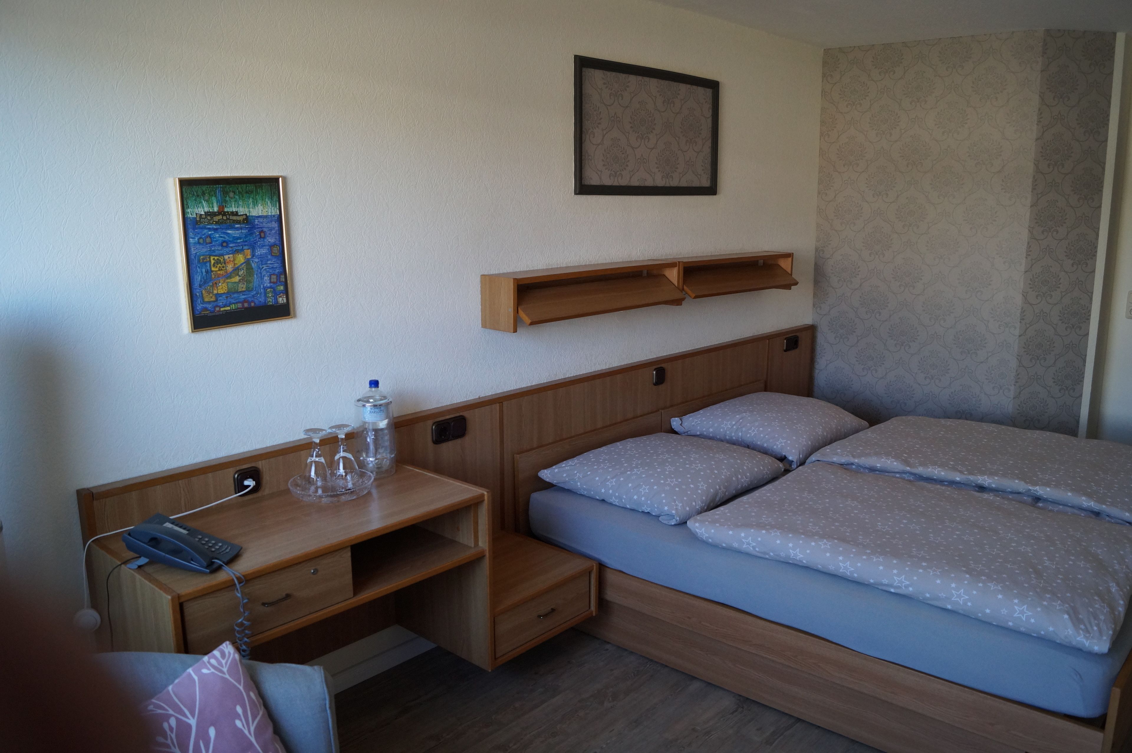 Een slaapkamer met een bed, een bureau met een telefoon, een waterfles en een lijst op de muur.
